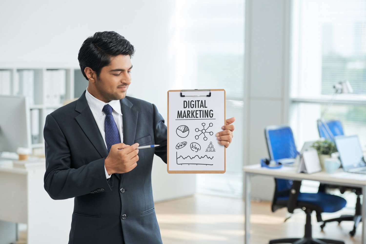 business man in suit holding clipboard and pointing at digital marketing poster in office